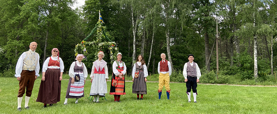 Deltagare från Väsby Folkdansgille står i folkdräkter framför en midsommarstång. 