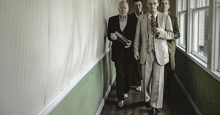 Fyra män i kostym går i en trång korridor, de håller munspel i händerna.