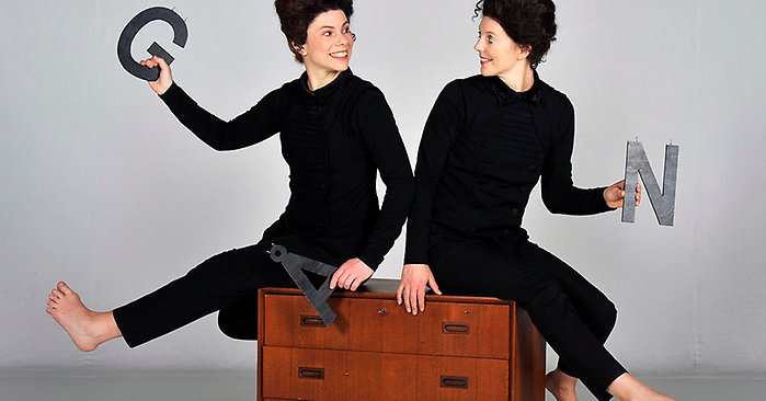 Två svartklädda personer med svarta peruker sitter på en brun byrå. De håller stora svarta bokstäver i händerna: G, Å och N.