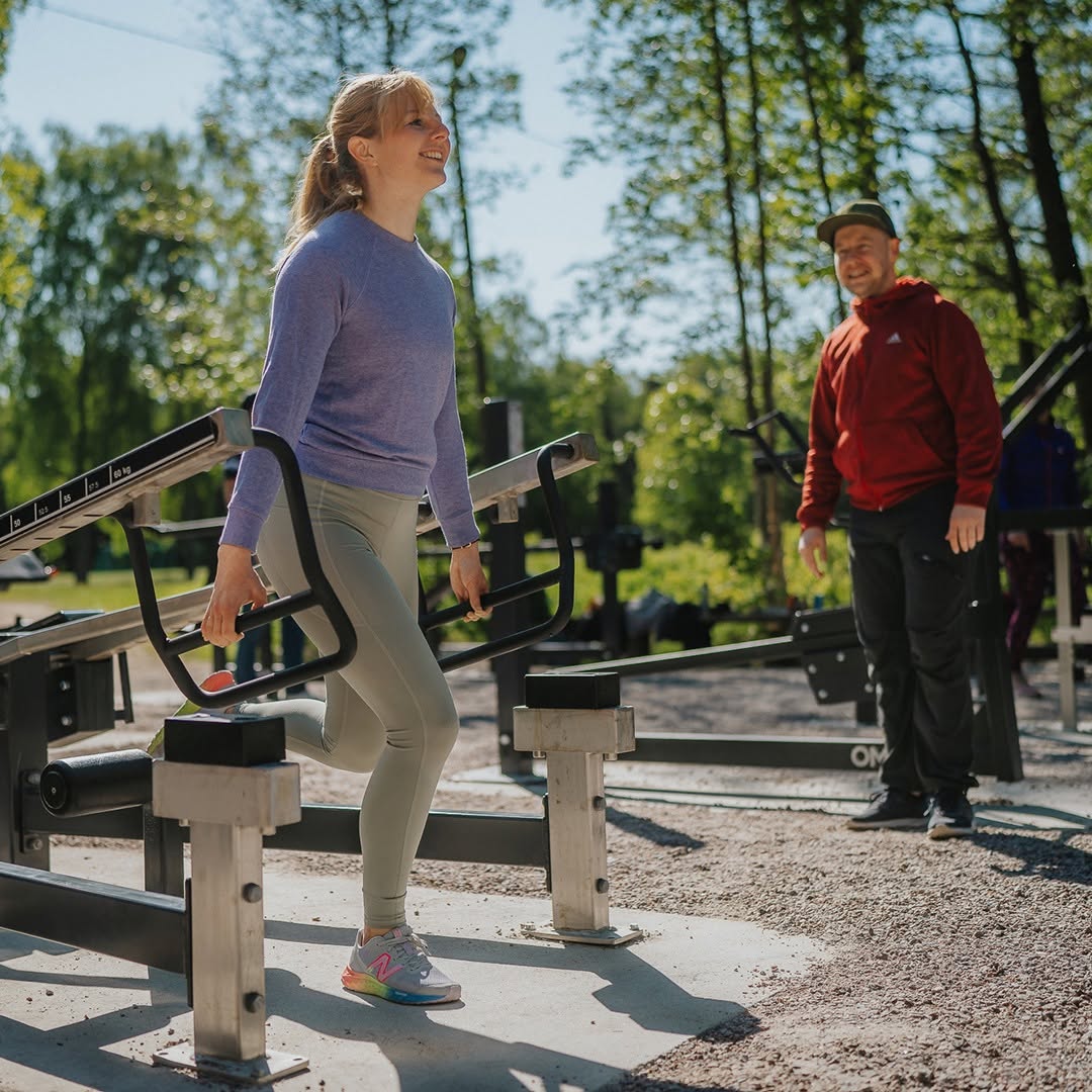 En kvinna tränar i ett utegym och gör en övning i maskin. I bakgrunden står en man och tittar på. Solig parkmiljö med träd.