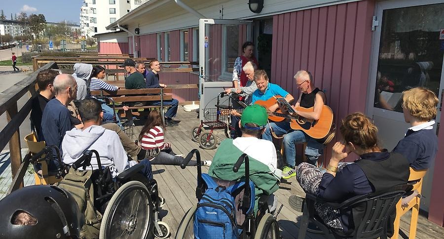 Musikstund på altanen. En man spelar på gitarr och i en ring framför honom sitter människor och lyssnar. Några av de som lyssnar sitter i rullstol.