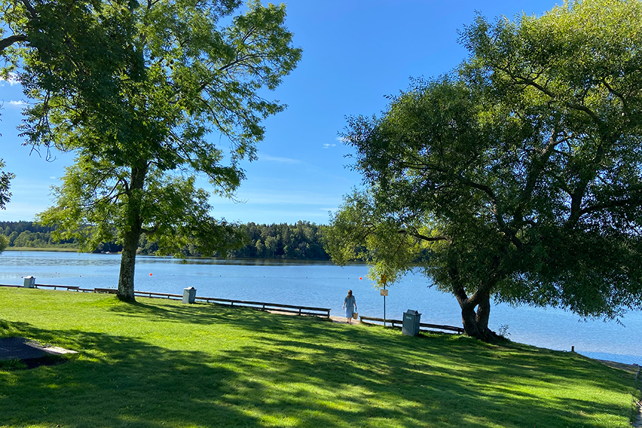 På bild syns en del av Kairobadet som är en av Mälarens badsjöar. På bild syns grönt gräs, blå himmel och en person som går på stranden.