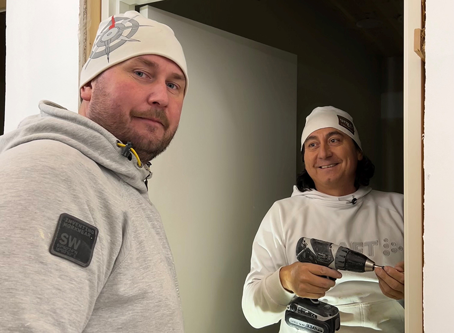 Två byggjobbare står inomhus vid en dörr och arbetar med renovering.
