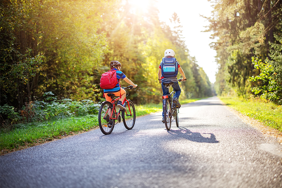 Två skolbarn cyklar i solskenet en sommardag med grönska på båda sidorna av vägen.