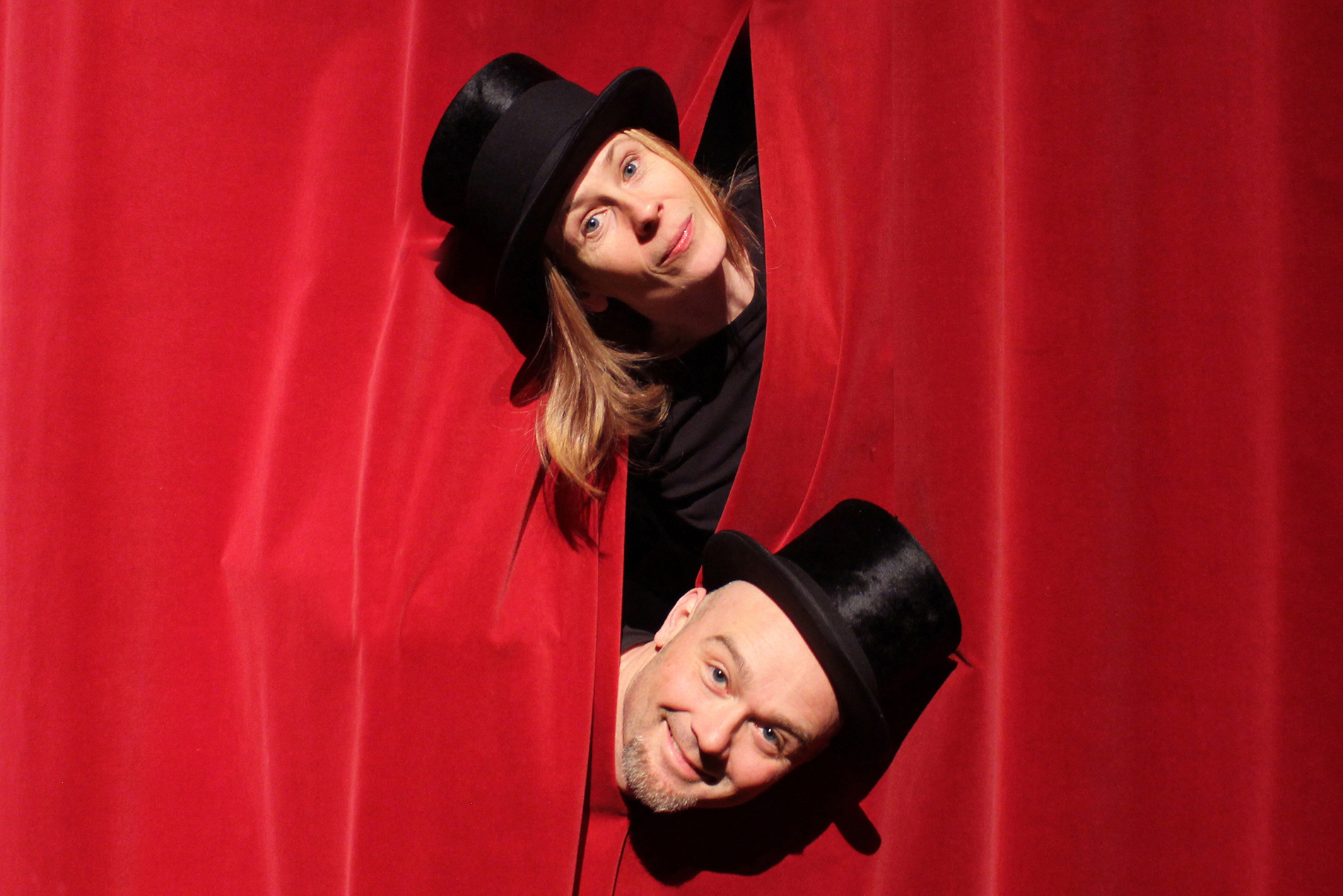 En man och kvinna med en svart hatt som tittar fram bakom en röd ridå.