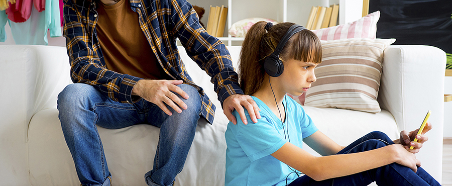 En vuxen person sitter i en vit soffa och har lagt handen på sin dotters axel. Dottern sitter med hörlurar på golvet och kollar på sin mobiltelefon. 