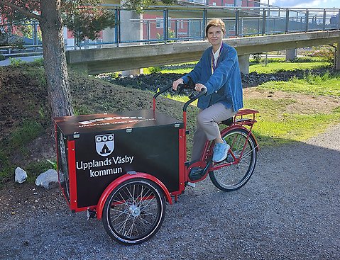 En kvinna sitter på en cykel med en låda som går att frakta saker i. Bokcykeln i Upplands Väsby, för att göra biblioteket tillgängligt för fler Väsbybor.