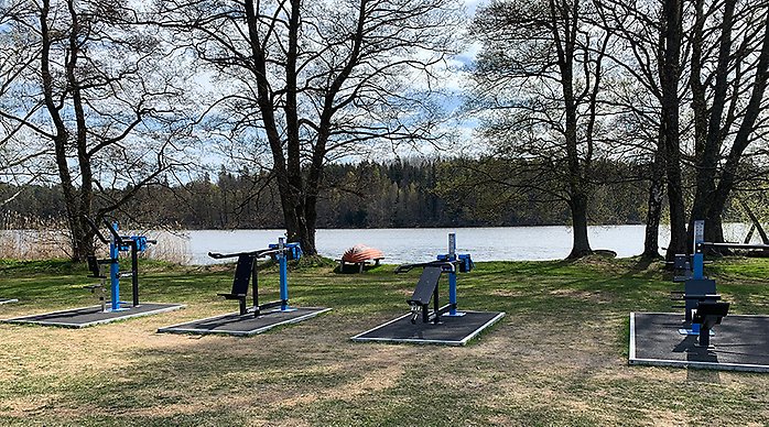 Olika träningsredskap i ett utegym med träd och vatten i bakgrunden.