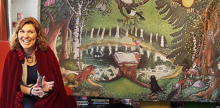 En kvinna i röd mantel berättar en saga framför ett tyg med bild av en sagoskog.