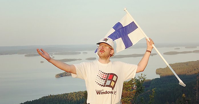 En man i en Windows-95-tröja viftar med finska flaggan framför ett vackert sjölandskap