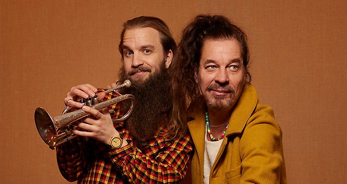 Två långhåriga män håller om varandra och den ena har en trumpet i handen