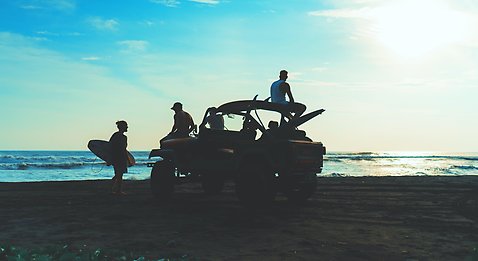 Några ungdomar som hänger på en strand. Några står på stranden och några sitter i sin terrängbil utan tak. Flera har sina surfingbrädor med sig. rad på stranden. 