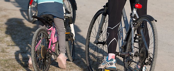 Man ser benen på ett barn som står med en cykel och bredvid ser man ett par vuxna ben intill en cykel. 