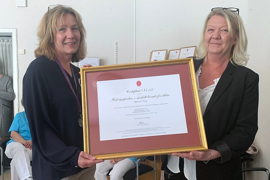 Två kvinnor håller upp ett certifikat med guldram och röd bakgrund
