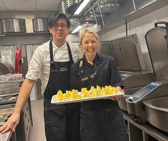 Två personer håller upp ett fat med gula desserter. De står i ett kök.