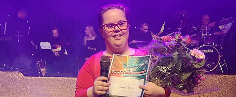 Josefin står på scenen och håller en mikrofon i handen tillsammans med ett diplom och en bukett blommor.