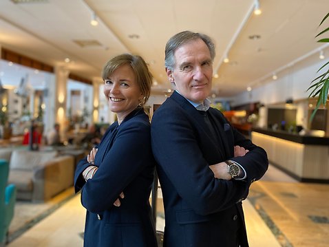 En kvinna och man står med ryggarna mot varandra och korsade armar och ler mot kameran.