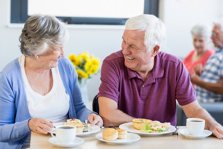 Två äldre personer skrattar och samtalar under en måltid.