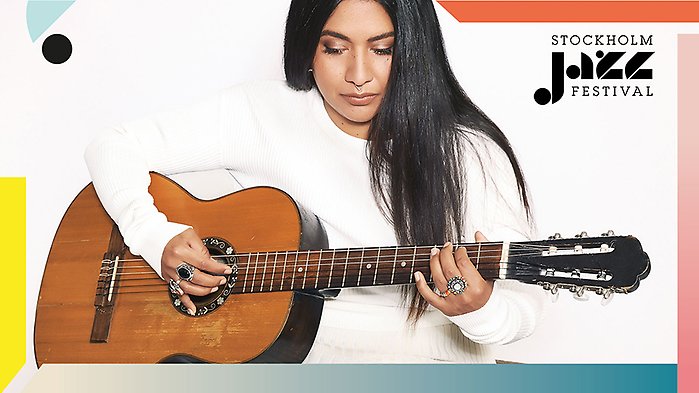 Kristin Amparo, i vit tröja och långt utsläppt mörkt hår, sittande med en akustisk gitarr, mot vik bakgrund