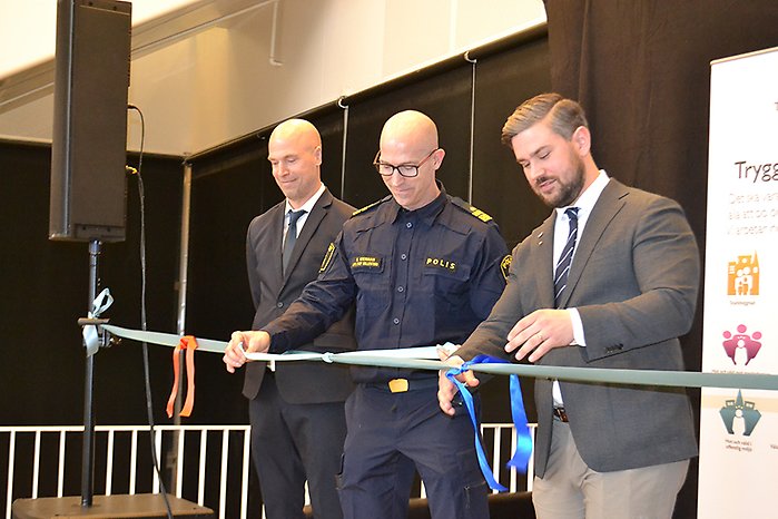 Tre men står på en scen och knyter ett varsitt mindre band på ett långt band.